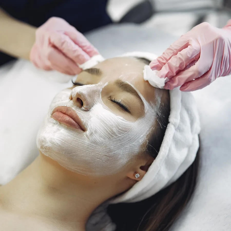 Profissional de luvas retira com algodão a máscara facial de uma cliente.