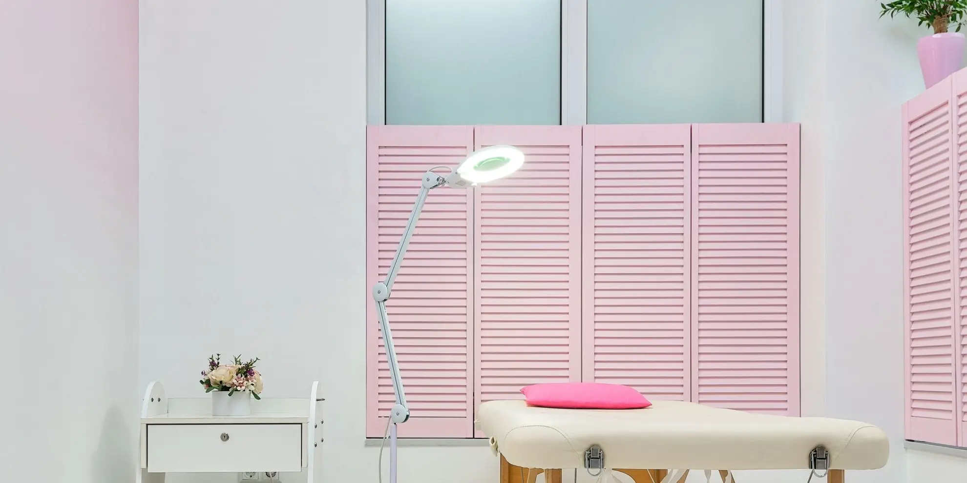 Sala de atendimento clara, com maca, luminária com lupa e venezianas rosa.
