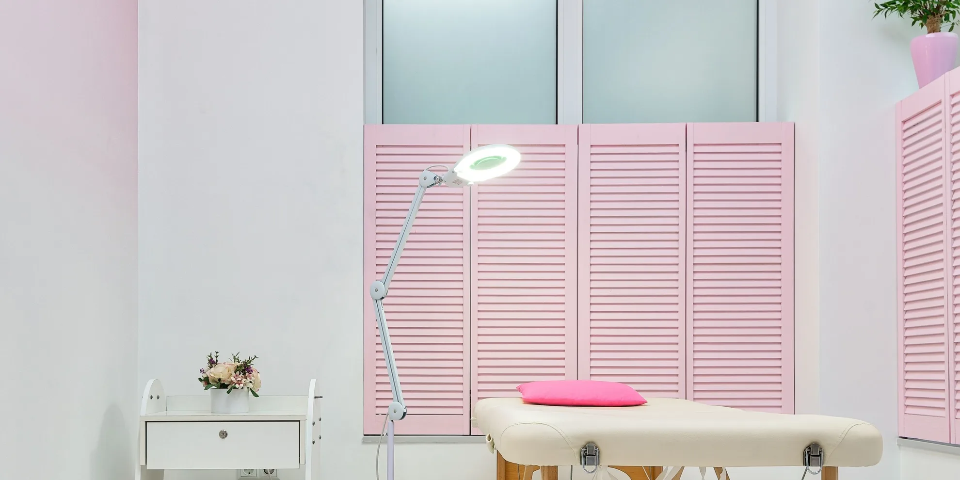 Sala de atendimento clara, com maca, luminária com lupa e venezianas rosa.