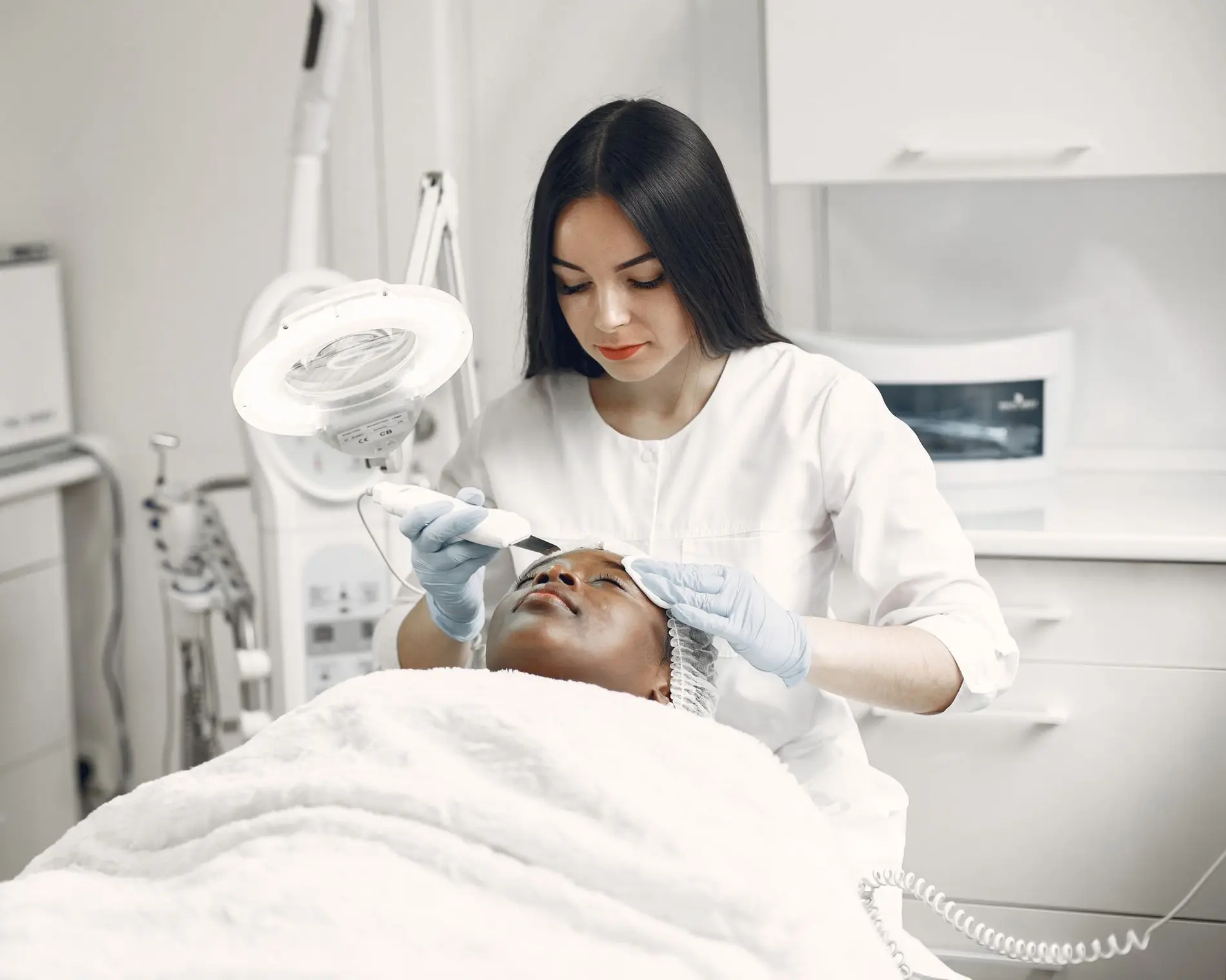 Esteticista de jaleco branco e luvas faz um tratamento facial em uma cliente deitada na maca.