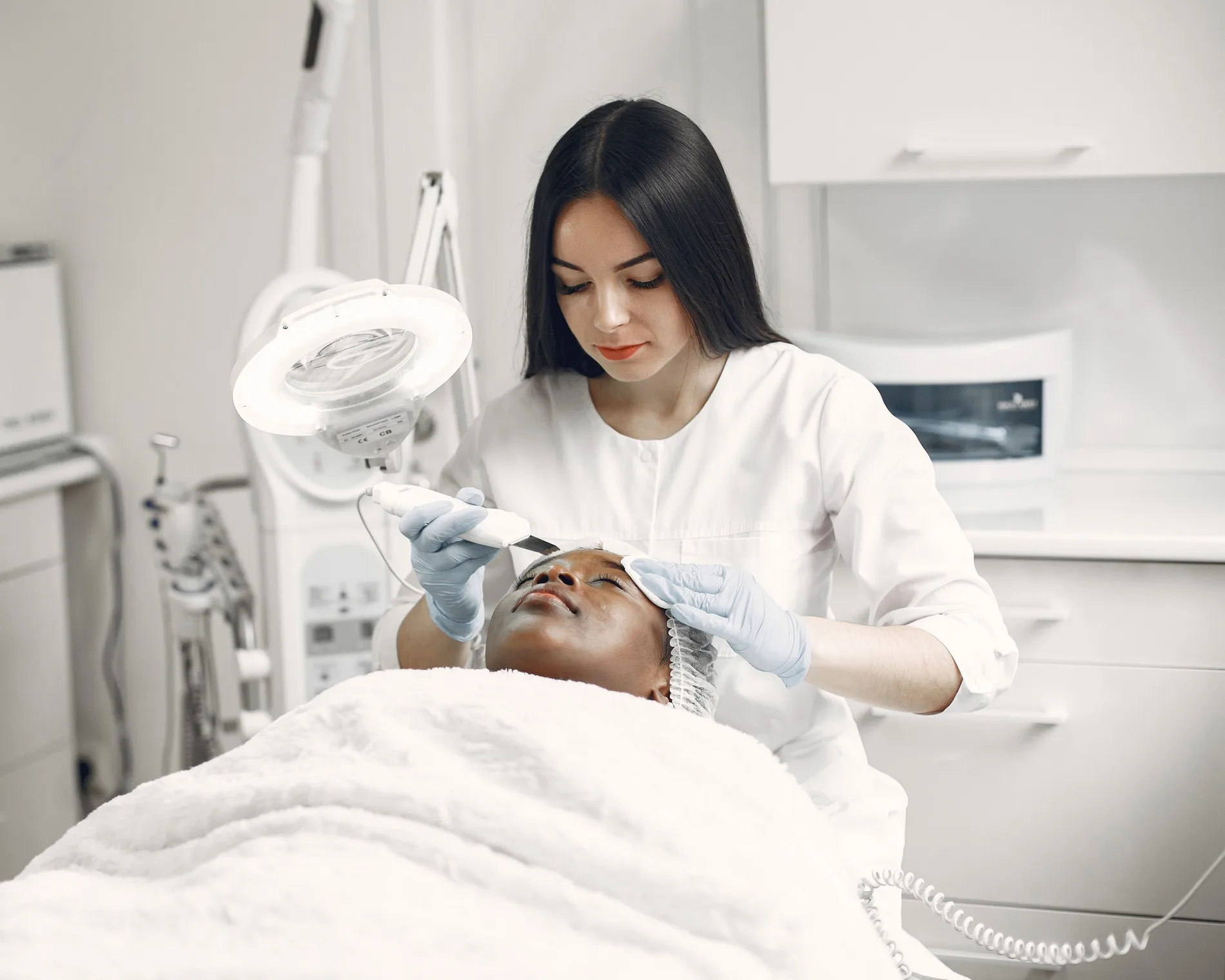 Esteticista de jaleco branco e luvas faz um tratamento facial em uma cliente deitada na maca.